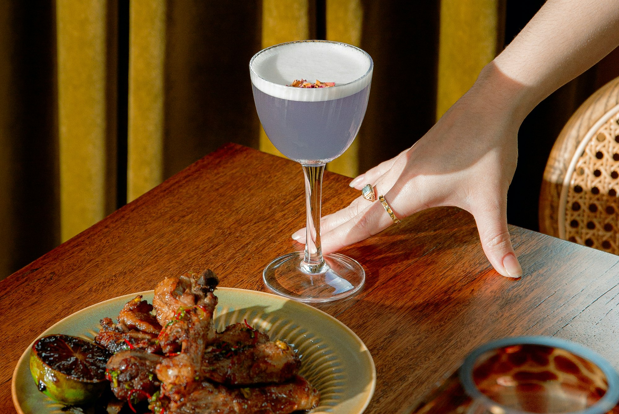 A purple cocktail and food sit on a timber table, with a hand resting next to them and olive green curtain in the background at Maggie May, Gasworks